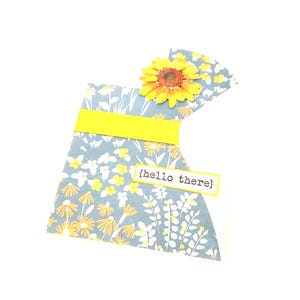 May include: A handmade greeting card with a floral design. The card features a yellow sunflower, a yellow band, and a blue floral pattern. The text "hello there" is printed on a small white rectangle.