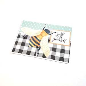May include: A greeting card featuring a large bee illustration with yellow and black stripes, and white wings. The card has a black and white checkered pattern at the bottom and a light blue polka dot pattern at the top. The text "just bee yourself" is written on the card.