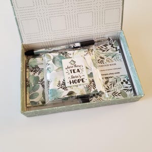 May include: A green and white gift box with a floral design. The box contains a small notebook, a pen, and a card that says "Where there's tea there's hope".