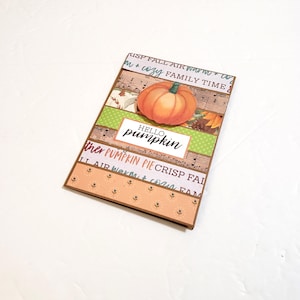 May include: A handmade greeting card with a 3D orange pumpkin and the words "Hello Pumpkin." The card features patterned paper with fall-themed text and designs, including musical notes and leaves. The card is rectangular with a brown border.