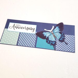 May include: A navy blue anniversary card with a large blue and black butterfly. The card has a white rectangle with the words "Happy Anniversary" in cursive. The card has sections with polka dots, stripes, and a checkered pattern.