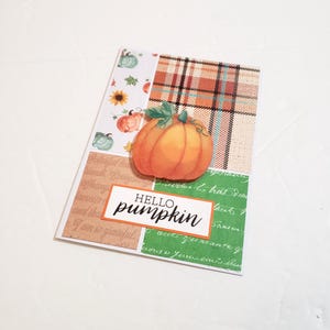 May include: A greeting card with a collage of patterns and a 3D orange pumpkin. The card has a plaid section, a section with pumpkins and sunflowers, and a section with green script. The words "HELLO pumpkin" are in a white rectangle.