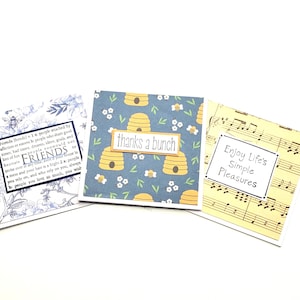 May include: Three square greeting cards. One card has a blue floral design with the word "FRIENDS" and a definition. Another card has a blue background with yellow beehives and the text "thanks a bunch". The third card has musical notes and the text "Enjoy Life's Simple Pleasures".