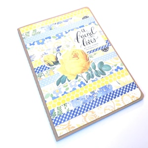 May include: A rectangular greeting card featuring horizontal strips of patterned paper in yellow, blue, and white. The design includes a large yellow rose illustration and the text A friend loves at all times Proverbs 17:17 in black script.
