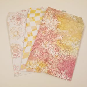 May include: Three decorative envelopes with different patterns. One envelope has a white background with a pink floral pattern. Another envelope has a white and yellow checkered pattern. The third envelope has a yellow background with a pink floral pattern.