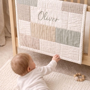 以下が含まれることがあります： 木製のベビーベッドに飾られた、名前「Oliver」が刺繍されたベビーキルト。キルトは白、ベージュ、グレーのニュートラルな色調のパッチワークデザインです。ぬいぐるみウサギがベビーベッドの中にいます。