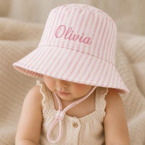 May include: A light pink and white striped bucket hat featuring the name Olivia embroidered in pink script on the front. The hat includes a matching pink and white striped chin strap with an adjustable toggle to secure the fit around the head.