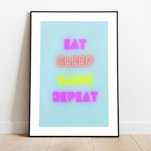 Könnte beinhalten: Ein gerahmter Druck mit hellblauem Hintergrund und neonrosa und gelbem Text, der "Essen Schlafen Spielen Wiederholen" lautet.