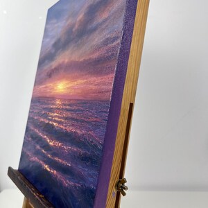 May include: A canvas painting of a sunset over the ocean. The artwork features a vibrant color palette of purples, pinks, and oranges, with the sun setting over the water. The canvas is mounted on a wooden easel.