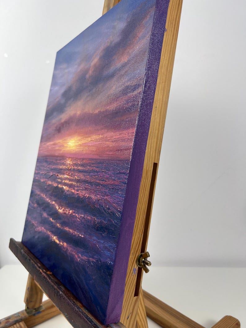 May include: A canvas painting of a sunset over the ocean. The artwork features a vibrant color palette of purples, pinks, and oranges, with the sun setting over the water. The canvas is mounted on a wooden easel.