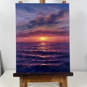 May include: An oil painting of a sunset over the ocean. The sky transitions from deep blue to shades of pink and orange, with clouds. The water reflects the sunset's light, creating shimmering waves. The artwork is displayed on a wooden easel.