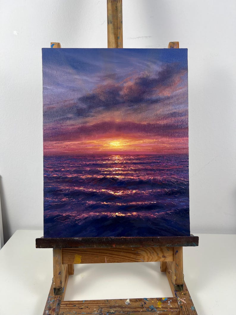 May include: An oil painting of a sunset over the ocean. The sky transitions from deep blue to shades of pink and orange, with clouds. The water reflects the sunset's light, creating shimmering waves. The artwork is displayed on a wooden easel.