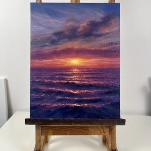 May include: An oil painting of a sunset over the ocean. The sky transitions from deep blue to shades of purple and orange, with clouds. The water reflects the sunset's colors, creating a shimmering effect. The painting is displayed on a wooden easel.