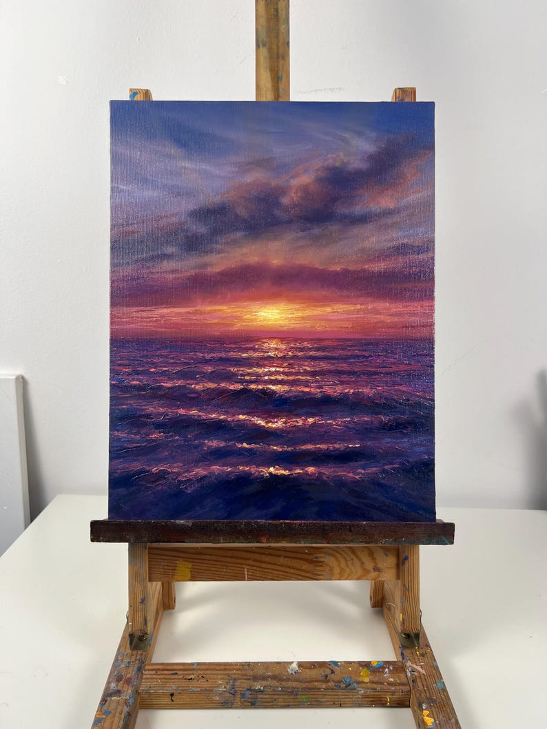 May include: An oil painting of a sunset over the ocean. The sky transitions from deep blue to shades of purple and orange, with clouds. The water reflects the sunset's colors, creating a shimmering effect. The painting is displayed on a wooden easel.