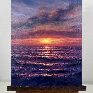 May include: Oil painting of a sunset over the ocean. The sky transitions from blue to purple and pink, with clouds. The sun is a bright yellow orb. The ocean is dark blue with white-capped waves. The painting is on a canvas on a wooden easel.