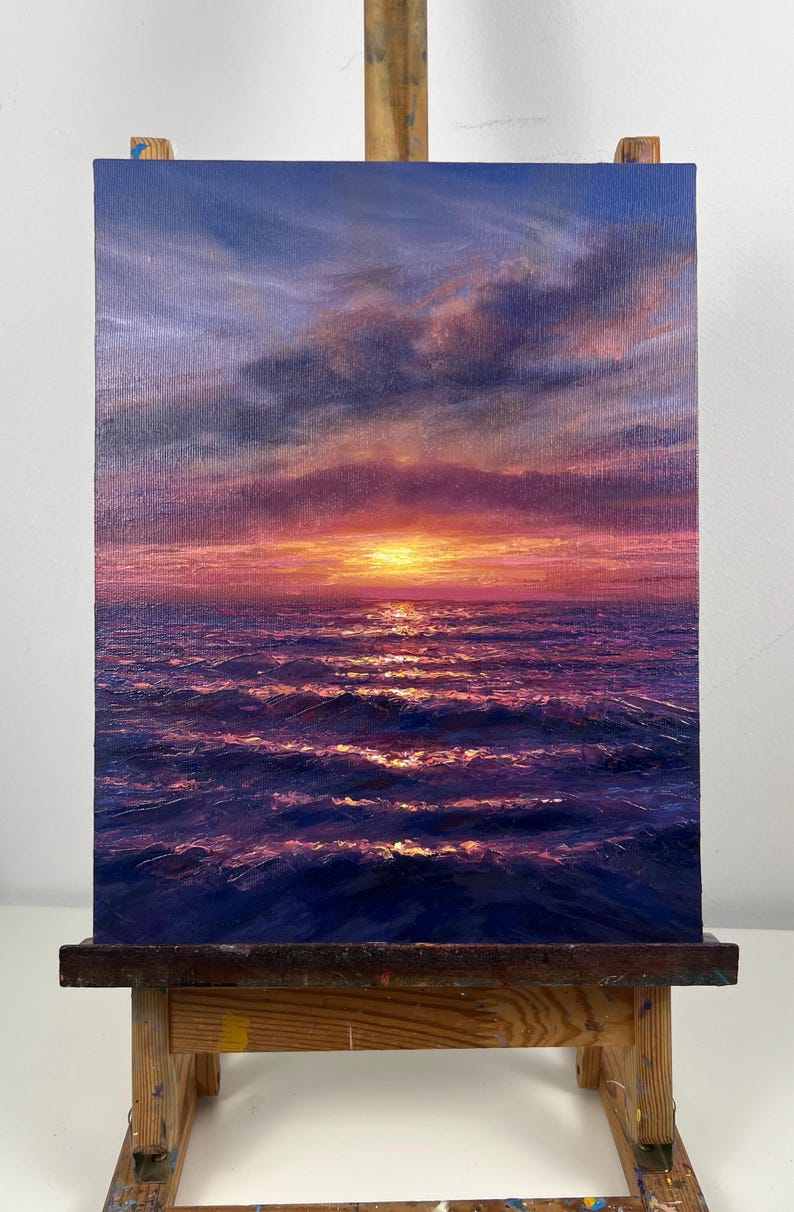 May include: Oil painting of a sunset over the ocean. The sky transitions from blue to purple and pink, with clouds. The sun is a bright yellow orb. The ocean is dark blue with white-capped waves. The painting is on a canvas on a wooden easel.