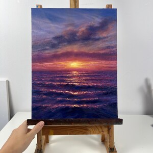 May include: An oil painting on canvas depicts a sunset over the ocean. The sky transitions from blue to pink and purple, with clouds. The sun is a bright yellow orb. The ocean waves reflect the colors of the sky. The painting is displayed on a wooden easel.