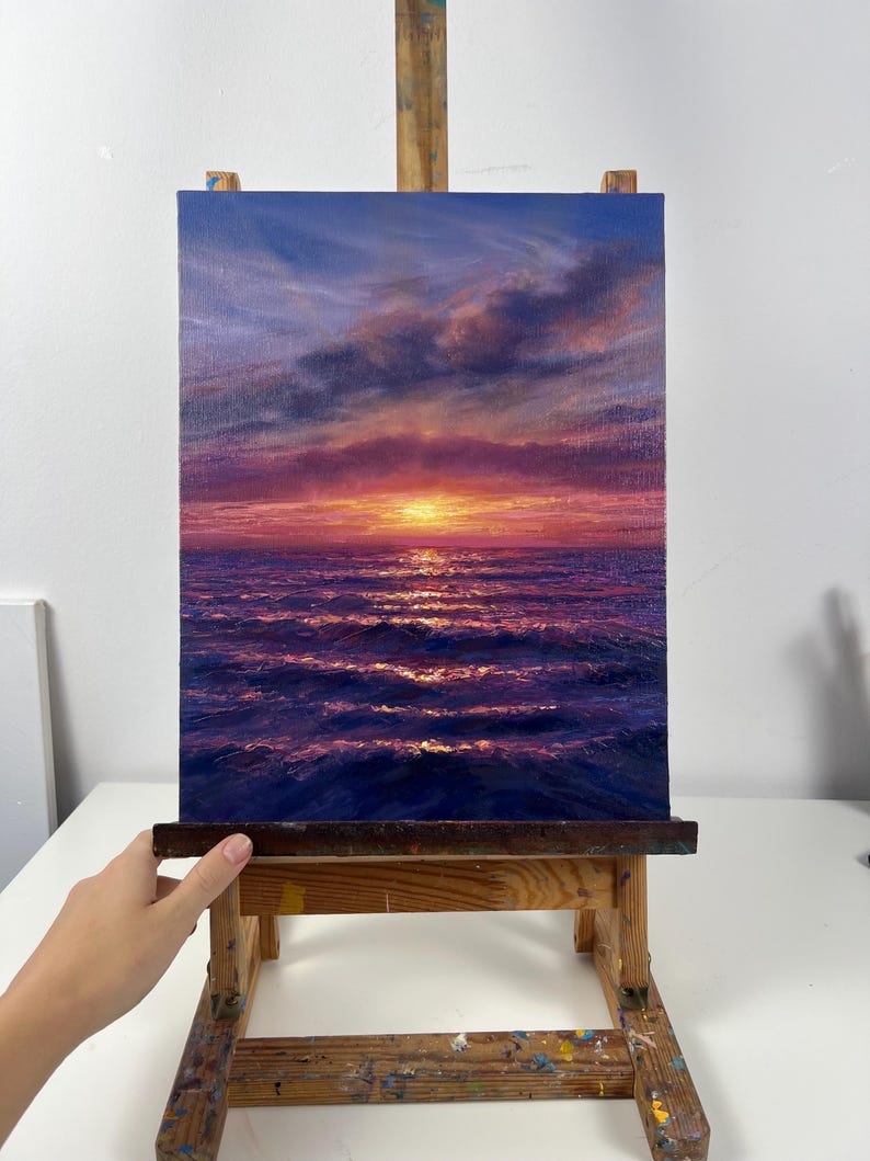 May include: An oil painting on canvas depicts a sunset over the ocean. The sky transitions from blue to pink and purple, with clouds. The sun is a bright yellow orb. The ocean waves reflect the colors of the sky. The painting is displayed on a wooden easel.