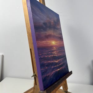 May include: An original oil painting of a sunset over the ocean, displayed on a wooden easel. The artwork features a vibrant color palette of purples, oranges, and blues, capturing the light reflecting on the water. The canvas has a purple border.