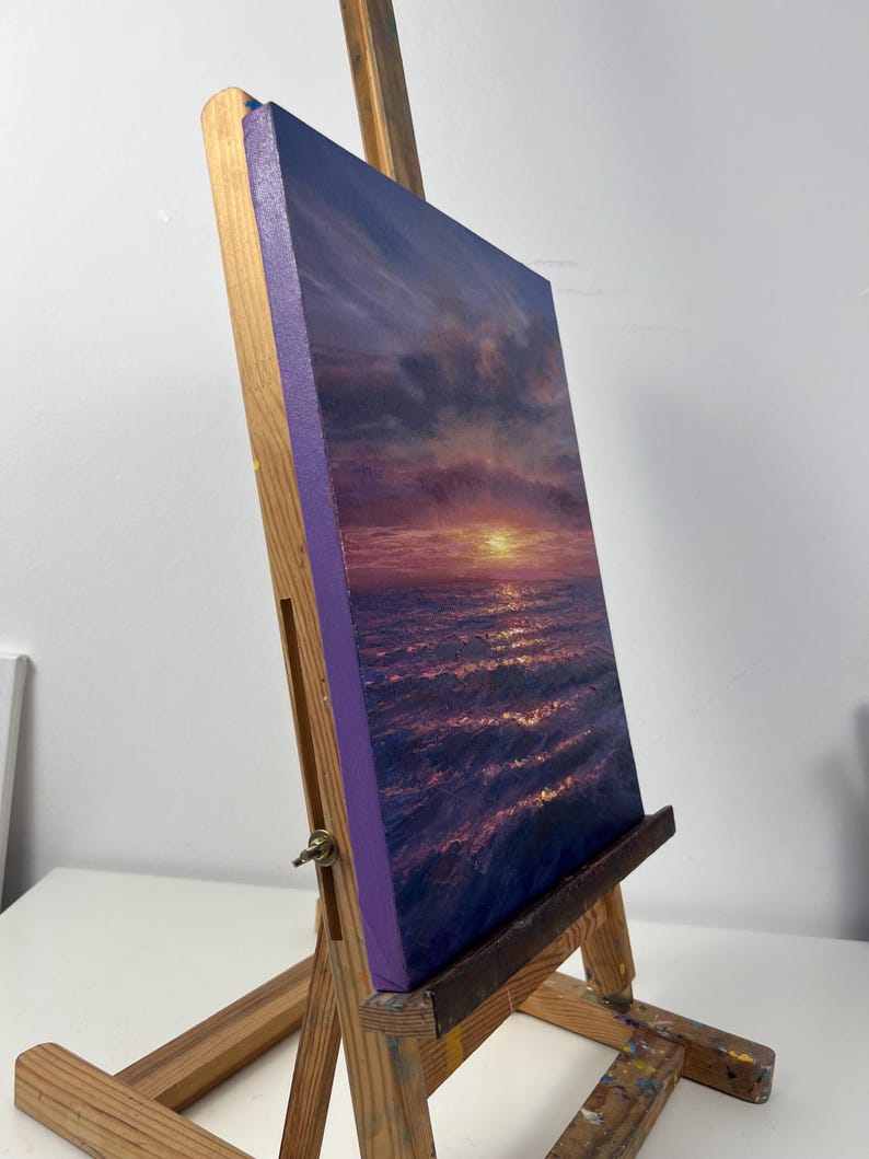 May include: An original oil painting of a sunset over the ocean, displayed on a wooden easel. The artwork features a vibrant color palette of purples, oranges, and blues, capturing the light reflecting on the water. The canvas has a purple border.