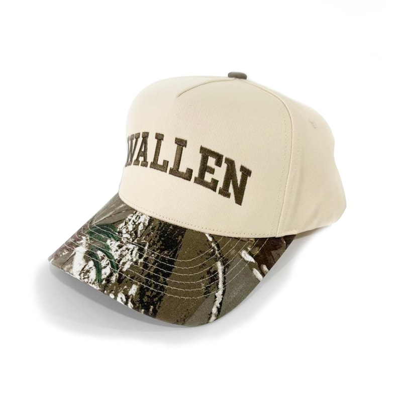 May include: A beige baseball cap with the word "WALLEN" embroidered in brown. The brim features a camouflage pattern in shades of green, brown, and white. The cap has a rounded crown and a curved brim.