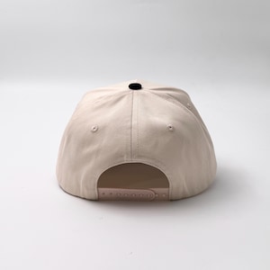 May include: A beige baseball cap with a curved brim and a black button on top. The back features an adjustable strap for a custom fit. The hat is made of a durable fabric and is suitable for casual wear.