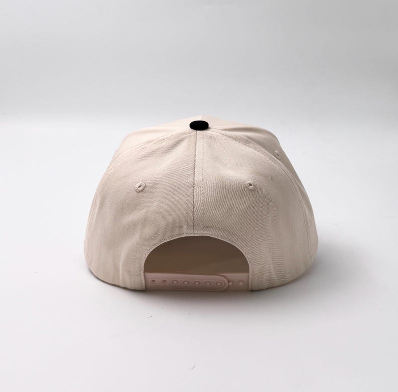 May include: A beige baseball cap with a curved brim and a black button on top. The back features an adjustable strap for a custom fit. The hat is made of a durable fabric and is suitable for casual wear.