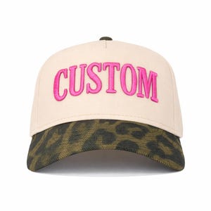 May include: A beige baseball cap with the word "CUSTOM" embroidered in bright pink across the front. The brim features a green and brown animal print, and the top button is dark green.