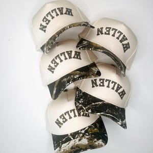 May include: A collection of six baseball caps. The caps are off-white with a camouflage pattern on the brim. The word "WALLEN" is embroidered in black on the front of each cap.