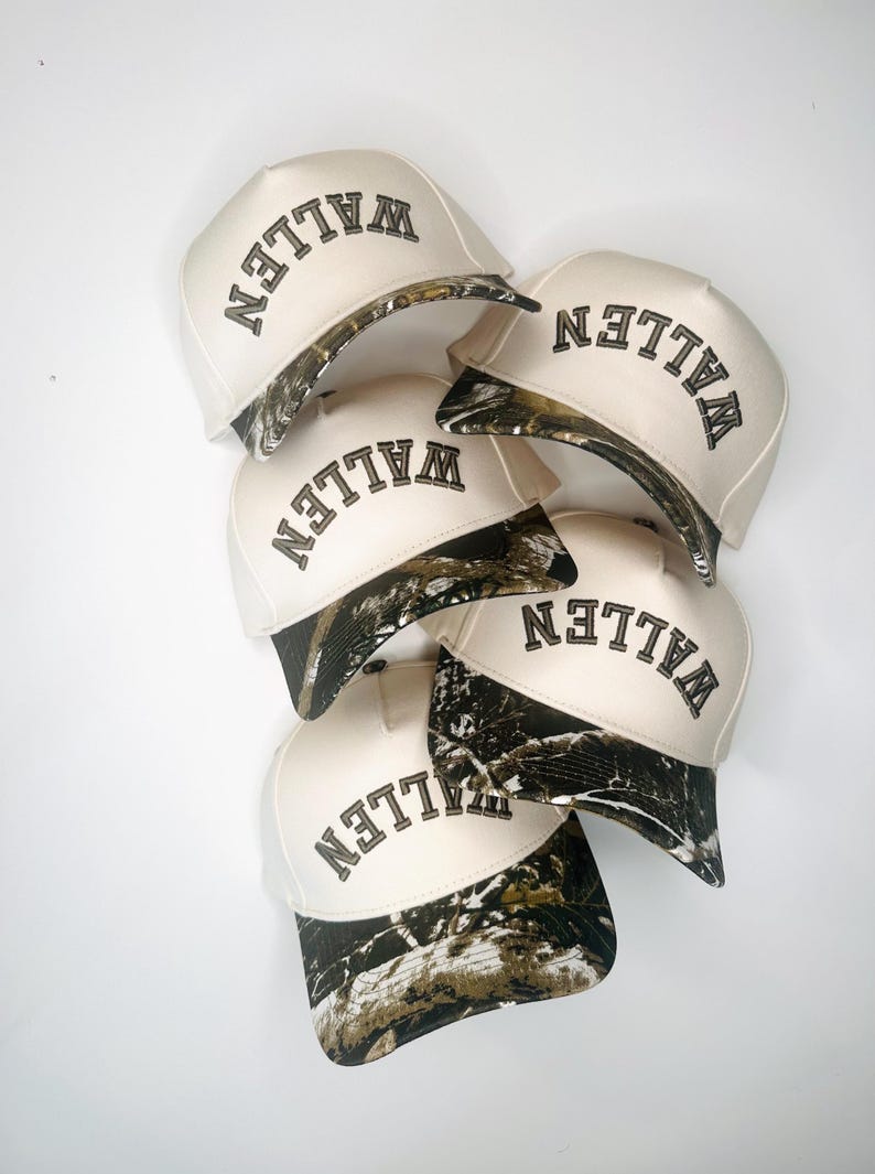 May include: A collection of six baseball caps. The caps are off-white with a camouflage pattern on the brim. The word "WALLEN" is embroidered in black on the front of each cap.