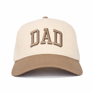 Puede incluir: Una gorra de béisbol de color crema con la palabra DAD bordada en letras marrones de bloque en el panel frontal. La gorra tiene una visera marrón sólida con costuras visibles y un botón marrón a juego en la parte superior de la corona.