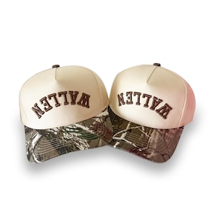 May include: Two baseball caps with a cream-colored crown and a camouflage brim. The word "WALLEN" is embroidered in brown across the front of each cap. The caps are set against a white background.
