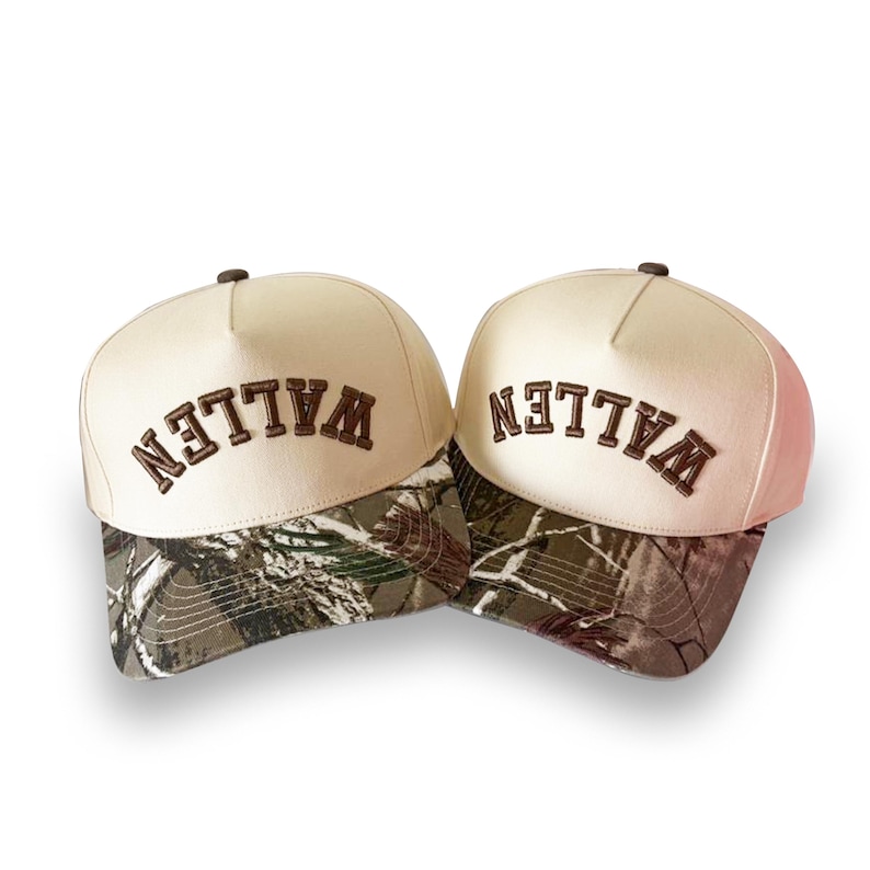 May include: Two baseball caps with a cream-colored crown and a camouflage brim. The word "WALLEN" is embroidered in brown across the front of each cap. The caps are set against a white background.