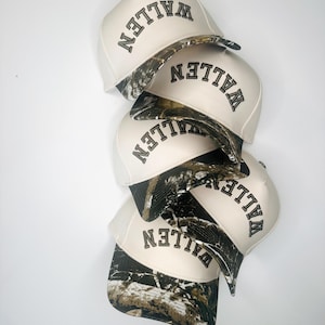 May include: A stack of cream-colored baseball caps with the word "WALLEN" embroidered in black. The brim of each cap features a camouflage pattern. The caps are stacked on a white background.