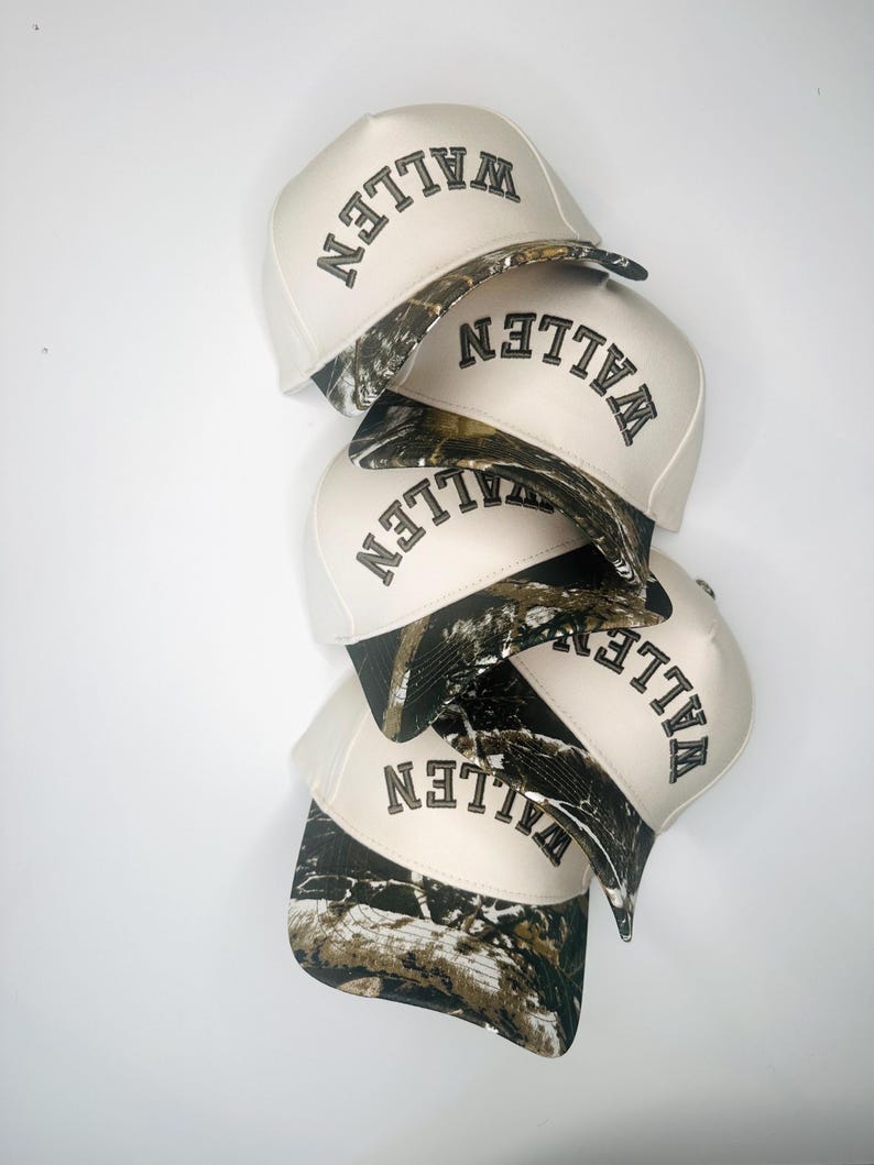 May include: A stack of cream-colored baseball caps with the word "WALLEN" embroidered in black. The brim of each cap features a camouflage pattern. The caps are stacked on a white background.