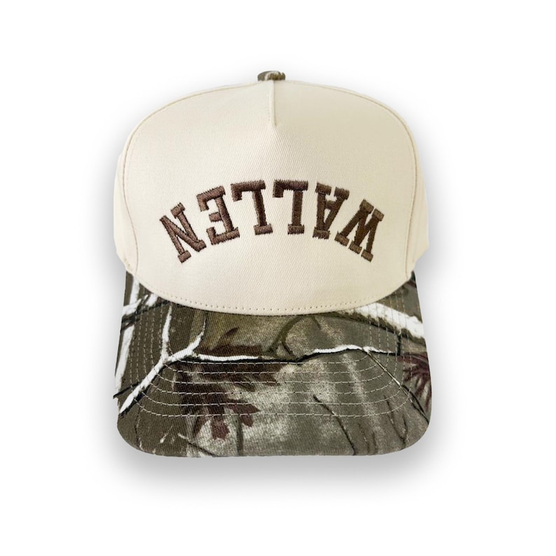 May include: A beige baseball cap with the word "WALLEN" embroidered in brown across the front. The brim features a camouflage pattern in shades of green, brown, and white. The cap has a structured crown and a curved brim.
