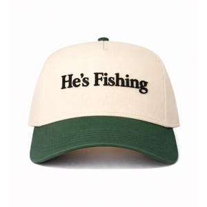 May include: A cream-colored baseball cap with a dark green brim. The phrase "He's Fishing" is embroidered in black across the front. The cap has a curved brim and a small green button on top.