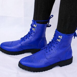 Könnte beinhalten: Ein Paar leuchtend blaue Lederstiefel mit Lyralochung und schwarzen Sohlen. Die Stiefel haben Schnürsenkel und silberfarbene Metallbeschläge. Die Stiefel werden mit schwarzen Socken getragen.