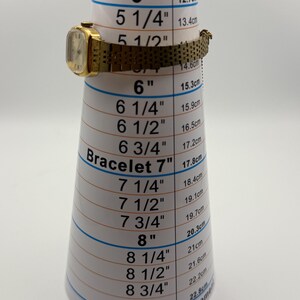 May include: A gold-toned vintage wristwatch with a rectangular face and a mesh band is displayed on a bracelet measuring cone. The cone shows measurements in inches and centimeters. The watch is approximately 5.5 inches (14 cm) in circumference.
