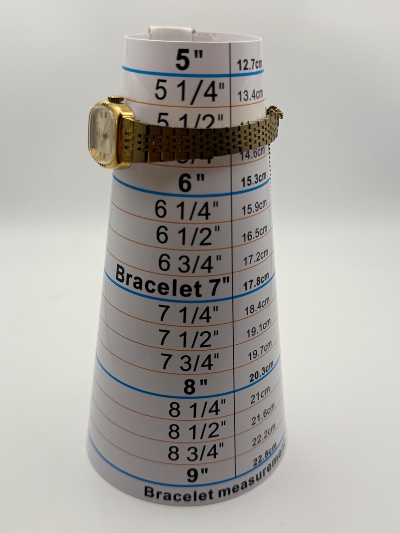 May include: A gold-toned vintage wristwatch with a rectangular face and a mesh band is displayed on a bracelet measuring cone. The cone shows measurements in inches and centimeters. The watch is approximately 5.5 inches (14 cm) in circumference.