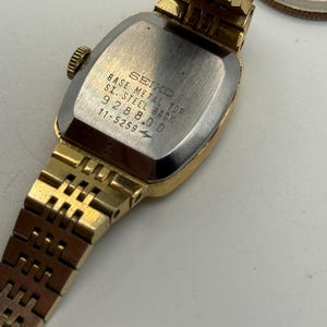 May include: A gold-toned Seiko wristwatch with a rectangular face and a matching gold band. The watch's back is silver with the text "SEIKO BASE METAL TOP S. STEEL BACK 928800 11-5259". The watch is partially open, revealing the inner workings.