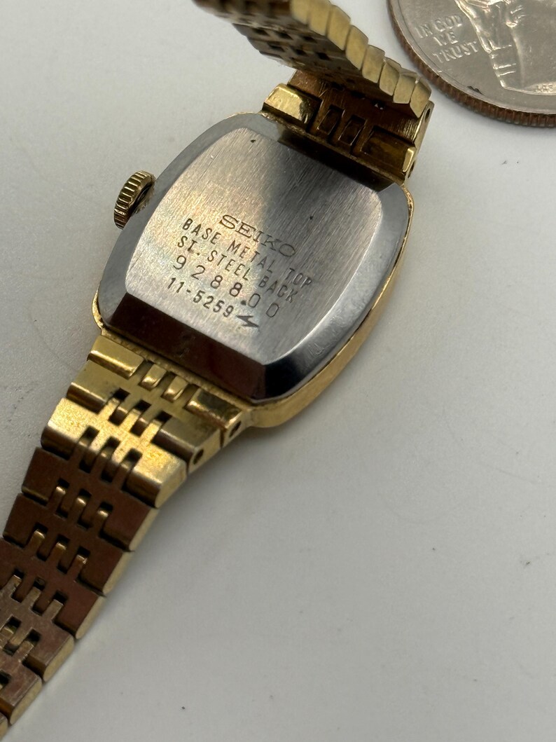 May include: A gold-toned Seiko wristwatch with a rectangular face and a matching gold band. The watch's back is silver with the text "SEIKO BASE METAL TOP S. STEEL BACK 928800 11-5259". The watch is partially open, revealing the inner workings.