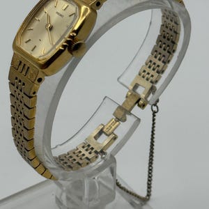 May include: A gold-toned Seiko wristwatch with a rectangular face and a matching linked band. The watch features a gold dial with simple hour markers and hands. The band is connected with a small chain.