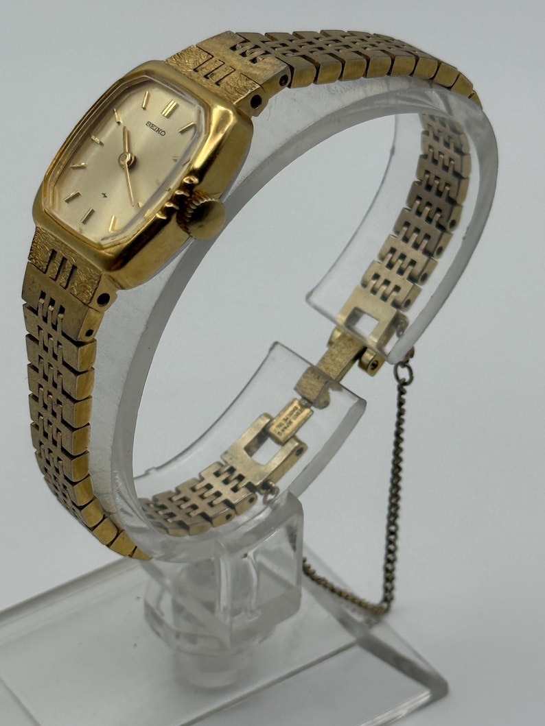 May include: A gold-toned Seiko wristwatch with a rectangular face and a matching linked band. The watch features a gold dial with simple hour markers and hands. The band is connected with a small chain.