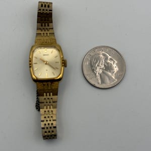 May include: A vintage gold-toned Seiko wristwatch with a rectangular face and a linked band. The watch is next to a quarter coin for size comparison. The watch face is a light cream color.