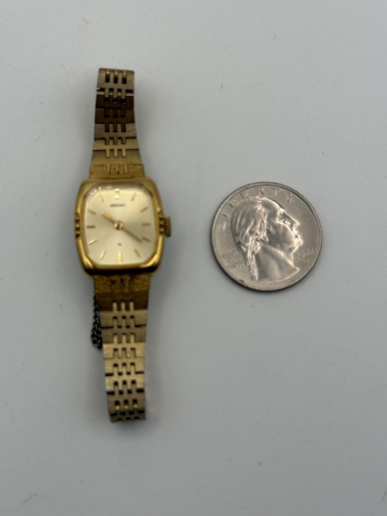 May include: A vintage gold-toned Seiko wristwatch with a rectangular face and a linked band. The watch is next to a quarter coin for size comparison. The watch face is a light cream color.