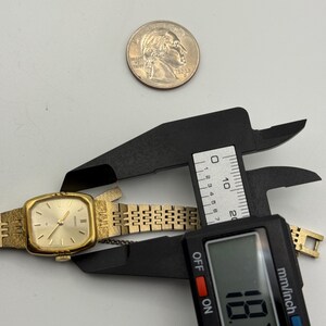 May include: A gold-toned Seiko quartz watch with a rectangular face and a linked band. The watch is being measured with a digital caliper, showing a measurement of 18.7 mm. A quarter coin is also present.