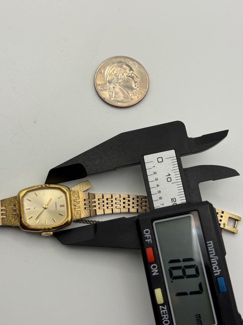 May include: A gold-toned Seiko quartz watch with a rectangular face and a linked band. The watch is being measured with a digital caliper, showing a measurement of 18.7 mm. A quarter coin is also present.