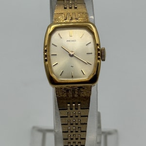 May include: A vintage gold-toned Seiko wristwatch with a rectangular face and a matching link band. The watch face is silver with black hour markers and hands. The watch is displayed on a clear stand.