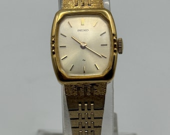 May include: A vintage gold-toned Seiko wristwatch with a rectangular face and a matching link band. The watch face is silver with black hour markers and hands. The watch is displayed on a clear stand.